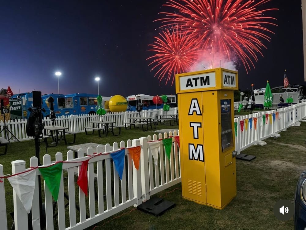 Yellow ATM Systems machine at an outdoor festival at night with fireworks overhead and colorful pennant flags